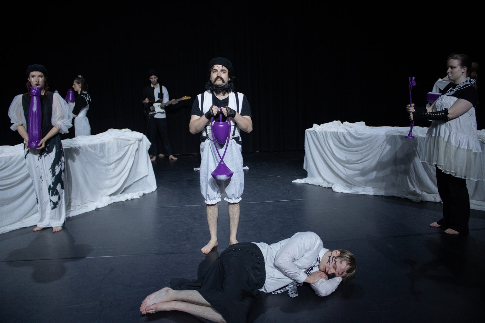 A scene from REM: performers in white costumes on a darkened stage holding purple props, with a guitarist playing upstage.